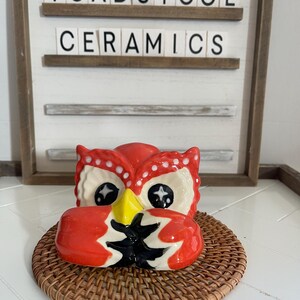 Può includere: Vaso da fiori a forma di gufo in ceramica rosso e bianco con dettagli neri e gialli. Il gufo ha un becco grande e occhi grandi con stelle bianche. Il vaso è appoggiato su un sottopentola in vimini marrone. Il cartello dietro il vaso recita "Toadstool Ceramics".