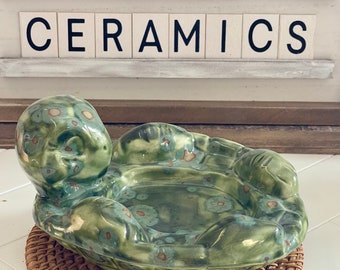 May include: Green ceramic turtle-shaped dish with a floral pattern. The dish is sitting on a woven brown mat. The text "TOADSTOOL CERAMICS" is visible in the background.