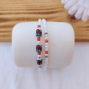 May include: Two beaded bracelets, one with white pearls and the other with red, white, and blue floral patterned beads.