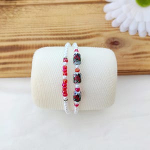 May include: Two beaded bracelets. One bracelet features white beads and red accent beads. The other bracelet has white beads and three rectangular beads with a red floral design. The bracelets are displayed on a white surface.