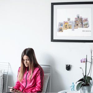 May include: A framed print of the floor plan of the apartment from the TV show "Friends". The print is hanging on a white wall above a couch with a person sitting on it. The person is looking down at a phone.