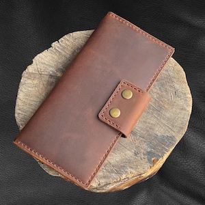 May include: A brown leather wallet with a flap closure and two brass poppers. The wallet is sitting on a wooden surface.