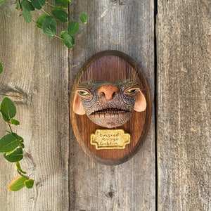 May include: A brown and grey goblin sculpture with a textured surface, mounted on a wooden plaque. The plaque has a gold label that reads "Crusted Arroyo Goblin".