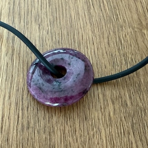 Può includere: Una collana con pendente rotondo con una pietra viola scuro e un cordino nero. La pietra, con un foro centrale, presenta sfumature rosa e viola. La collana è esposta su una superficie di legno.
