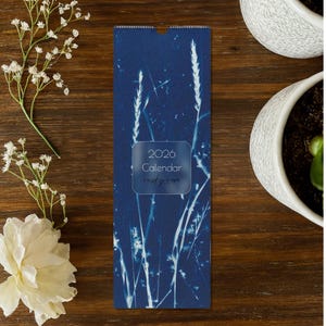 May include: A navy blue 2026 calendar with a botanical design featuring white plant silhouettes. The calendar has a rectangular shape with a rounded top and includes the text "2026 Calendar rusty rose". The calendar is on a wooden surface with white flowers and a potted plant.