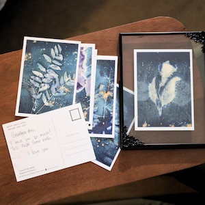 May include: A collection of rectangular postcards featuring dark blue botanical prints with gold accents, alongside a black metal frame containing a similar print. One postcard shows handwritten text: Grandma Bev, I miss you so much! Tell Papa Gene hello. I love you.