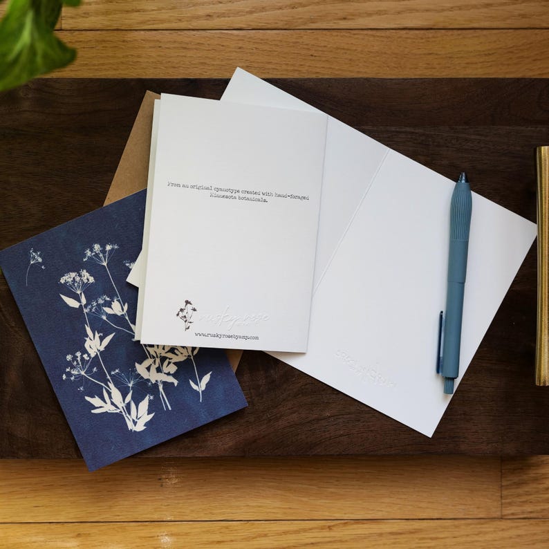 Puede incluir: Un conjunto de art&iacute;culos de papeler&iacute;a, que incluye una tarjeta azul marino con un dise&ntilde;o floral blanco, una tarjeta blanca con texto y un bol&iacute;grafo azul. Las tarjetas est&aacute;n sobre una superficie de madera oscura.
