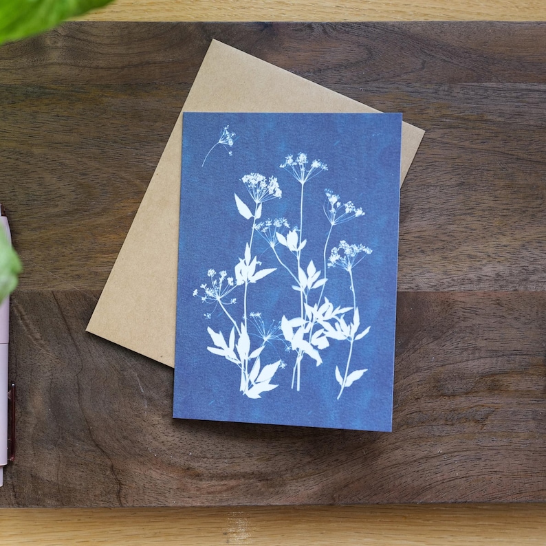 Puede incluir: Una tarjeta de felicitaci&oacute;n azul con un dise&ntilde;o bot&aacute;nico blanco de flores y hojas delicadas. La tarjeta se presenta con un sobre marr&oacute;n, sobre una superficie de madera.