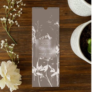 May include: A 2026 calendar with a floral design in white against a muted brown background. The text "2026 Calendar rusty rose" is displayed in a rectangular, transparent box. The calendar is displayed on a wooden surface with white flowers.