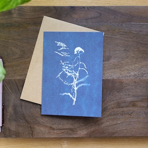 Puede incluir: Una tarjeta de felicitaci&oacute;n azul con una impresi&oacute;n bot&aacute;nica blanca de una planta con hojas delicadas y una flor. La tarjeta va acompa&ntilde;ada de un sobre marr&oacute;n. Los art&iacute;culos est&aacute;n sobre una superficie de madera.