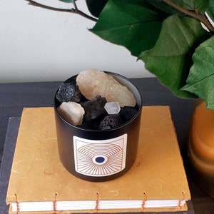 Puede incluir: Un difusor de cerámica negro con un diseño geométrico y un motivo de ojo blanco y azul. El difusor está lleno de rocas volcánicas negras y cristales blancos.