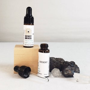 Op de afbeelding: Twee kleine bruine glazen flesjes Honey Brass geurverspreiderolie met zwarte druppelaars, één op een houten blok. De flesjes zijn gelabeld met de geur "Desert" en "FO". Er zijn ook zwarte stenen en heldere kristallen.