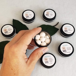 May include: A set of ten small, white, round, scented beads in a black metal container. The container has a label that says "HONEY BRASS" and "PRAIRIE" with a list of scents: cotton blossom, rosemary, spiced honey. The container is sitting on a white surface with other similar containers labeled with different scents.