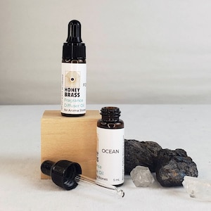 May include: Two small brown glass bottles with droppers, one labeled "Honey Brass Fragrance Diffuser Oil for Aroma Stones" and the other labeled "Ocean Fragrance Diffuser Oil for Aroma Stones" with a 5 ml volume. The bottles are on a wooden surface with black and white stones.
