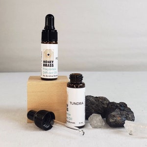 Puede incluir: Dos pequeños frascos de vidrio marrón con goteros, etiquetados como "Honey Brass" y "Tundra". Los frascos contienen aceite de difusor de fragancia para piedras aromáticas. Los frascos están sobre una superficie de madera con piedras negras y blancas.