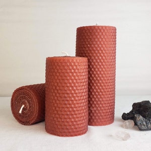 May include: Three red beeswax candles with a honeycomb pattern. The candles are different sizes and are arranged on a white surface.