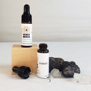 Op de afbeelding: Twee bruine glazen flesjes met druppelaars, een met het label "HONEY BRASS Fragrance Diffuser Oil for Aroma Stone" en de andere met het label "FOREST Fragrance Diffuser Oil 5 ml".