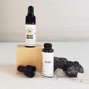 Puede incluir: Dos pequeños frascos de vidrio marrón con goteros, uno etiquetado como "Honey Brass Fragrance Diffuser Oil for Aroma Stones" y el otro como "River Fragrance Oil". Los frascos están sobre una superficie de madera con pequeñas piedras y cristales.