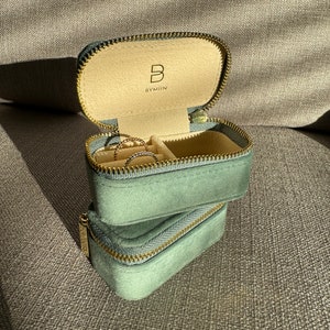 May include: A green velvet jewellery box with a gold zipper and the text "B BYMIIN" on the inside lid. The box is open and there are three rings inside.