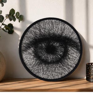 Könnte beinhalten: Runde Wandkunst mit einem schwarzen Augendesign aus Fadengrafik. Das Kunstwerk befindet sich vor einem weißen Hintergrund und ist mit einem schwarzen Rahmen versehen. Ein dekorativer Kerzenhalter und eine Vase mit Grün sind ebenfalls zu sehen.
