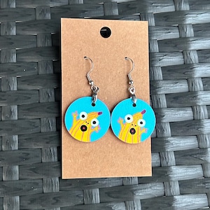 May include: A pair of round earrings with a cartoon monster design. The earrings have a light blue background with a yellow monster illustration. The earrings are attached to silver-coloured hooks and are displayed on a brown card.