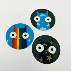 May include: Three round painted cat stickers with different backgrounds. One cat has a rainbow background, another has a blue sky background with clouds, and the last one has a black background with gold stars.