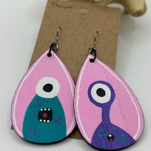 May include: A pair of pink teardrop-shaped earrings with painted monster faces. One monster is teal with a white eye and open mouth, the other is purple with a white eye and a small mouth. The earrings are made of wood and have titanium hooks.