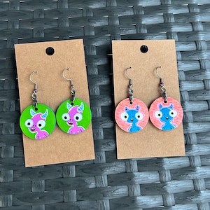 May include: Two pairs of handmade earrings. One pair features a pink monster on a green background, the other pair has a blue monster on a pink background. The earrings are round and made of wood, with silver-colored hooks.