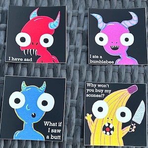 May include: Four square art prints with cartoon monster illustrations. The monsters are red, pink, blue, and yellow, each with large white eyes and horns. Text includes "I have sad," "I ate a bumblebee," "What if I saw a butt," and "Why won't you buy my scones!?"