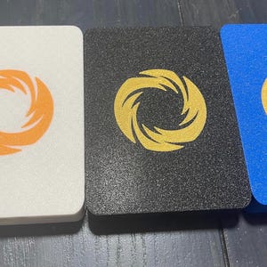 May include: Three rectangular coasters with a textured, granular surface. The left coaster is white with an orange spiral logo, the center is black with a gold spiral logo, and the right is blue with a gold spiral logo centered on each piece.