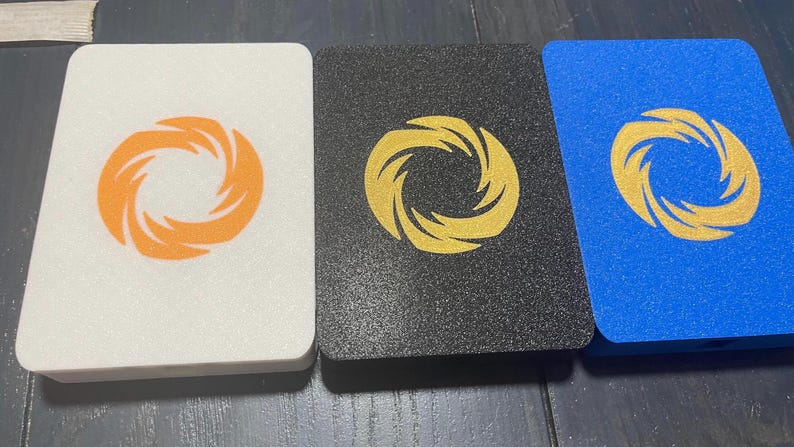 May include: Three rectangular coasters with a textured, granular surface. The left coaster is white with an orange spiral logo, the center is black with a gold spiral logo, and the right is blue with a gold spiral logo centered on each piece.