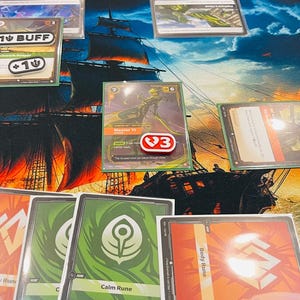 May include: A tabletop card game setup featuring various playing cards with green and red designs, including cards labeled Calm Rune and Body Rune. A central card displays a character illustration with a red token marked with a 3, and buff tokens.