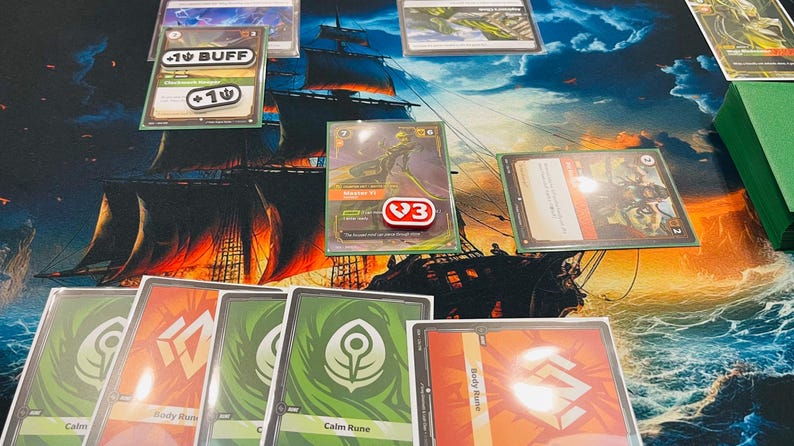 May include: A tabletop card game setup featuring various playing cards with green and red designs, including cards labeled Calm Rune and Body Rune. A central card displays a character illustration with a red token marked with a 3, and buff tokens.
