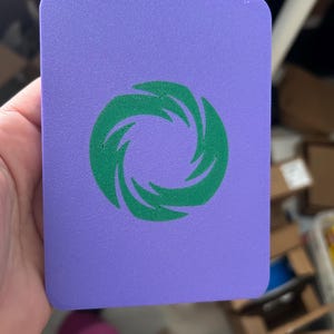 May include: A rectangular purple card with rounded corners featuring a centered green graphic of a stylized swirling vortex or circular flame pattern. The surface of the card has a fine, uniform texture throughout the entire purple and green area.
