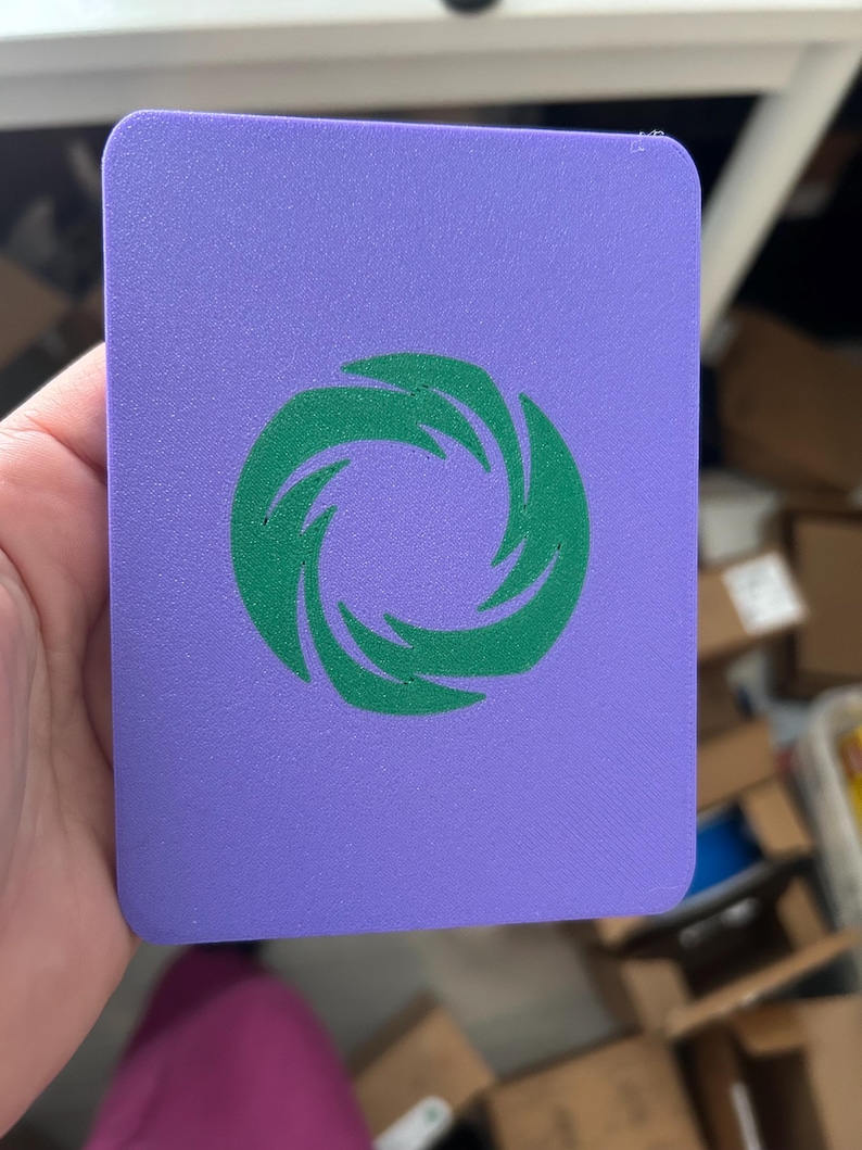 May include: A rectangular purple card with rounded corners featuring a centered green graphic of a stylized swirling vortex or circular flame pattern. The surface of the card has a fine, uniform texture throughout the entire purple and green area.