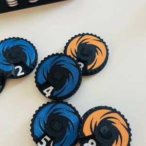 May include: Circular game tokens with black, blue, and orange designs. Each token has a number from 2 to 8. The tokens are on a white surface, with a black box in the background. The tokens have a spiral design.