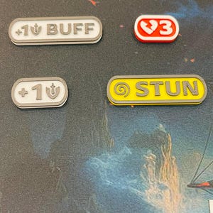 May include: Four rectangular game tokens with rounded corners. One white token reads +1 BUFF, one white token reads +1, one red token displays a heart icon with the number 3, and one yellow token reads STUN with a circular icon on the left side.