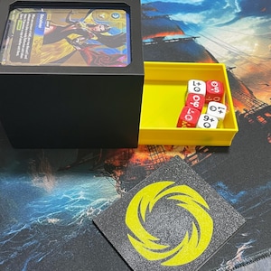 Könnte beinhalten: Eine schwarze Kartenaufbewahrungsbox mit einem gelben Würfelbehälter und einer schwarz-gelben Spielmatte. Die Box enthält illustrierte Karten. Die Würfel sind rot und weiß. Die Spielmatte hat ein gelbes Spiraldesign.