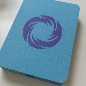 May include: A rectangular light blue plastic case featuring a centered purple graphic of a swirling vortex or spiral design. The surface has a textured finish, and a small rectangular port is visible on the bottom edge of the case.