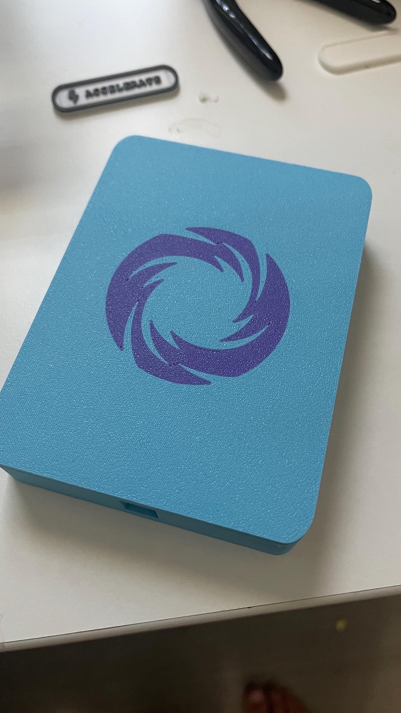 May include: A rectangular light blue plastic case featuring a centered purple graphic of a swirling vortex or spiral design. The surface has a textured finish, and a small rectangular port is visible on the bottom edge of the case.