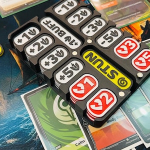 May include: A black and white game accessory with slots for tokens and cards. The accessory has the word "STUN" in yellow and white, and includes tokens with numbers and "BUFF" labels. Cards with text and graphics are visible.