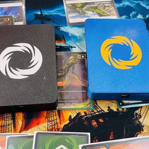May include: Two rectangular card deck boxes, one black with a white circular spiral emblem and one blue with a yellow circular spiral emblem, positioned on a game mat featuring a ship illustration and various trading cards scattered around them.