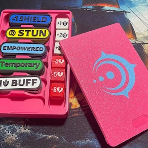 May include: A pink plastic game token organizer with a matching lid. The tray holds rectangular tokens labeled +1 SHIELD, STUN, EMPOWERED, Temporary, and +1 BUFF, alongside red and white dice marked with numerical values and heart or anchor icons.