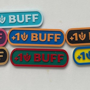 May include: A collection of seven rectangular plastic badges with rounded corners, each featuring the text +1 BUFF and a small sword icon. The badges come in various color combinations including light blue, purple, orange, black, red, green, and yellow.