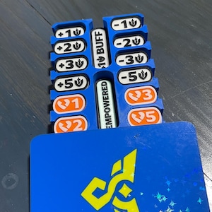 May include: A blue plastic game token organizer featuring two vertical columns of slots holding white and orange rectangular tiles with numerical values and symbols. The center divider displays the text BUFF and EMPOWERED. A blue card with a yellow logo sits in front.