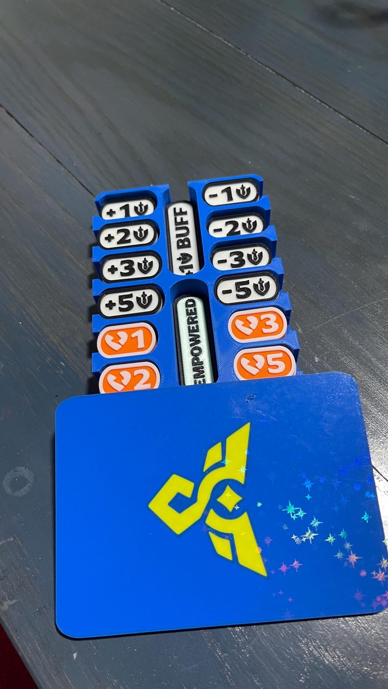 May include: A blue plastic game token organizer featuring two vertical columns of slots holding white and orange rectangular tiles with numerical values and symbols. The center divider displays the text BUFF and EMPOWERED. A blue card with a yellow logo sits in front.