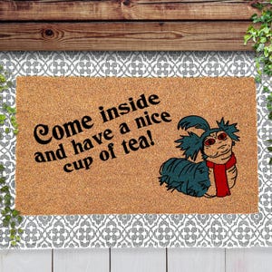 May include: A brown doormat with a white border and a white and gray patterned design. The doormat has the text "Come inside and have a nice cup of tea!" and a cartoon image of a blue and green worm with red scarf.