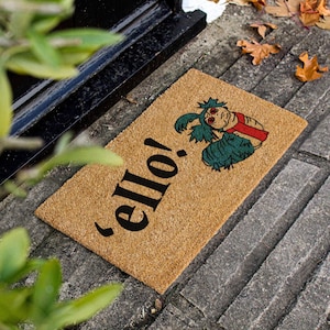 May include: A brown coir doormat with the text 'ello!' in black lettering and a green and red cartoon creature with large eyes.