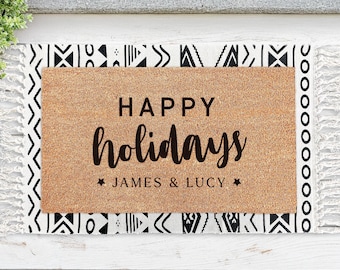 Puede incluir: Una alfombra de bienvenida marrón con el texto "Happy holidays" en letras negras cursivas. Debajo del texto se encuentra "James & Lucy" en letras negras. La alfombra tiene un borde con un patrón geométrico en blanco y negro.