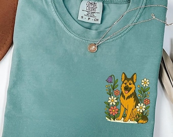 Puede incluir: Camiseta Comfort Colors de color verde azulado con un diseño de un perro pastor alemán rodeado de flores coloridas. La camiseta tiene una pequeña etiqueta en el cuello y un collar plateado con un colgante.